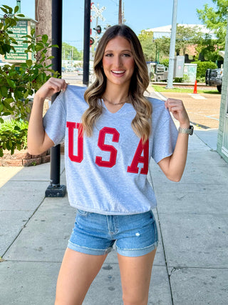 A person wearing a grey t-shirt with 'USA' printed in red letters, paired with blue denim shorts, standing on a sidewalk.