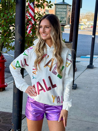 A woman wearing a white hoodie with the phrase 'YALL' sequin embellishments, paired with purple shorts, standing on a sidewalk.