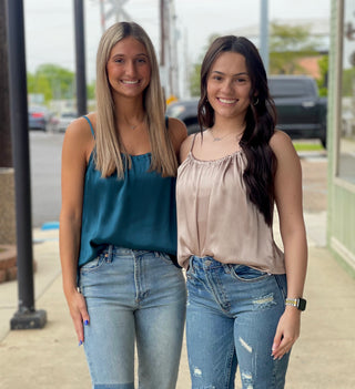 Two women standing on the sidewalk, smiling and posing in front of a store. One woman is wearing a green crinkle satin camisole with a round-neckline, and the other is wearing a beige top with blue jeans.