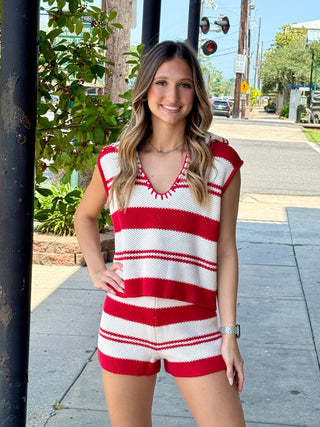 A female model wearing a sleeveless red and white striped sweater and shorts set standing on a sidewalk.