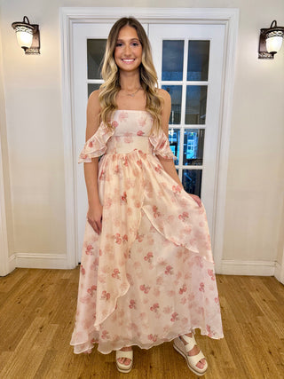 A woman wearing a pink and red floral print dress with off-shoulder sleeves, standing in front of a door with floral print.