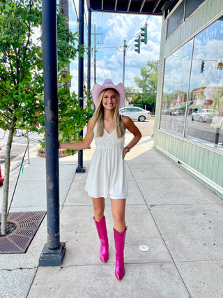 A woman standing on the sidewalk wearing a white romper with a pink cowboy hat and boots, with a storefront in the background.