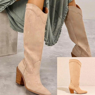 A pair of beige suede boots with a pointed toe and a low heel, displayed against a backdrop of a woman's dress and a flat sole view of the boots.
