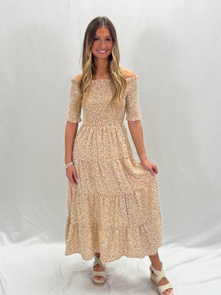 A woman standing and smiling, wearing an apricot dress with a floral print and a round neckline.