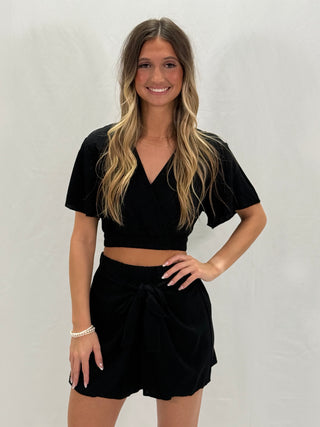 A woman wearing a black crop top with a cross knot detail and matching shorts.