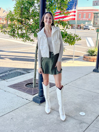 A woman standing on a sidewalk wearing a cream cropped puffer vest with drawstrings and pockets, paired with a green skirt and white boots.