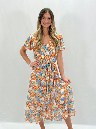 A woman standing and posing in a yellow floral ruffled dress with short sleeves and a V-neck neckline.