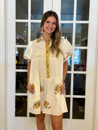 Woman wearing a white dress with gold footballs & accents in front of a glass door.