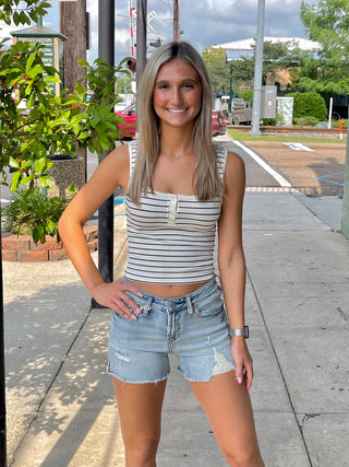 A woman standing on the sidewalk wearing a striped tank top and denim shorts.