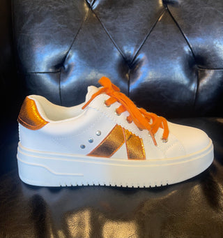 A pair of white tennis shoes with orange crinkle metallic accents and orange laces, placed on a black leather surface.