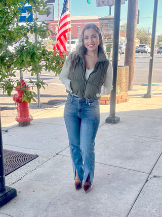 A person standing on a sidewalk wearing high-waisted, cropped straight-leg jeans and a green jacket with a vest.