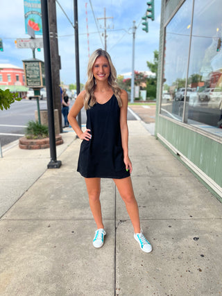 Woman standing on sidewalk wearing a black athletic cotton dress with white and blue metallic slip on sneakers.