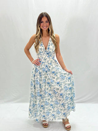 A woman standing and smiling, wearing a blue floral maxi dress with a V-neckline and sleeveless top.