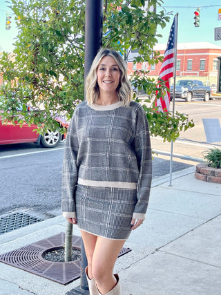 A woman standing on the sidewalk wearing a knee-length mini skirt with sleeves and a solid-color panel on the sleeves, paired with boots and a stone check sweater.
