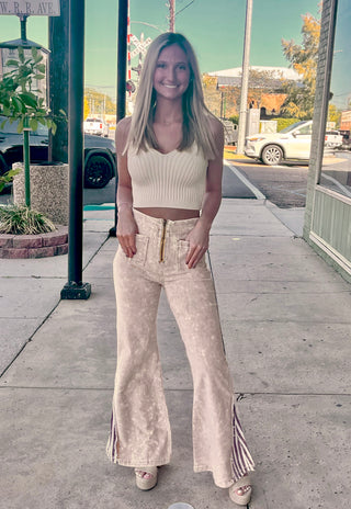 A woman standing on the sidewalk wearing cream spaghetti crop top with flare high rise yellow and striped denim pants.