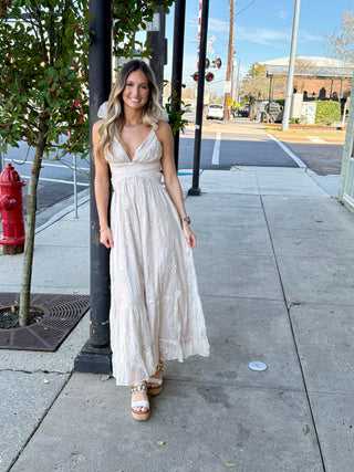 A woman standing on a sidewalk wearing a champagne white floral dress with sleeveless top and maxi length.