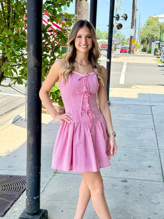 A woman standing on the sidewalk against a pole, wearing a pink sleeveless mini dress with a button-up front.