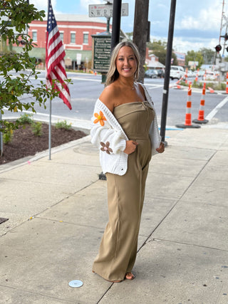 Woman in beige jumpsuit with floral cover-up standing on a sidewalk with traffic and American flag in background