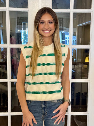 A person wearing a green and white striped sleeveless top paired with jeans, standing in front of a glass door.