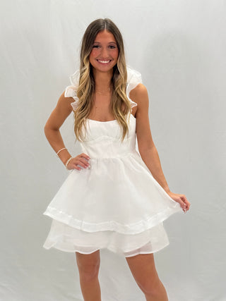 A woman wearing a white organza tiered mini dress with short sleeves and a square neckline.