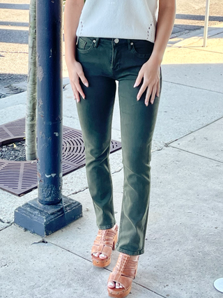 A person standing on the sidewalk wearing green mid-rise slim straight leg jeans with a white top and brown heel sandals.