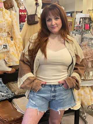 Woman standing in front of boutique scenery with a cream V-neck shirt on paired with cream and brown cardigan and blue distress jean shorts