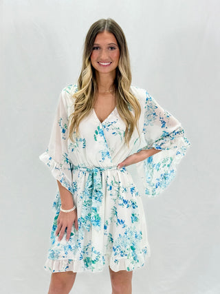 A woman wearing a white floral ruffle trim dress with 3/4 length sleeves, knee-length, with a v-neck neckline.
