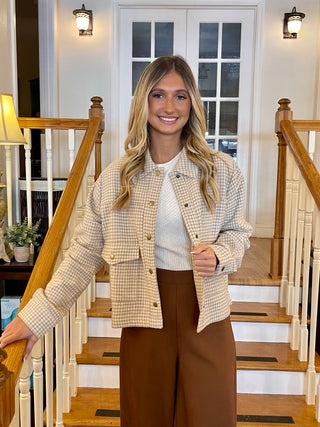A woman standing on a staircase wearing a taupe button crop jacket and brown trousers, with a checkered pattern on the jacket.