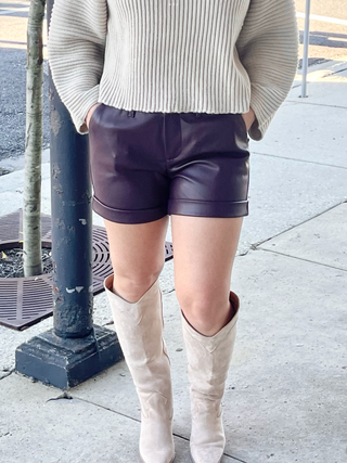 A pair of burgundy high-waisted shorts with a cuffed hem, paired with a cream-colored, long-sleeve top and beige boots.