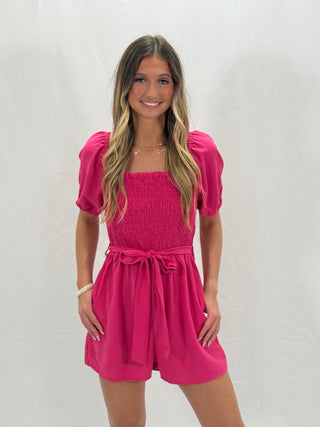 A woman wearing a pink solid smocked romper with short puffed sleeves and a bow at the waist.