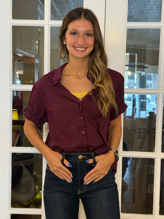 Woman in a plum shirt and jeans standing in front of a window.