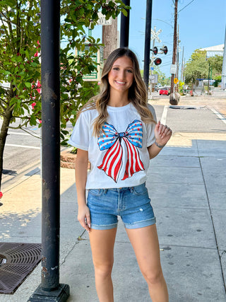 A woman standing on the sidewalk, wearing a white t-shirt with a red and blue stripe bow design, and blue denim shorts.
