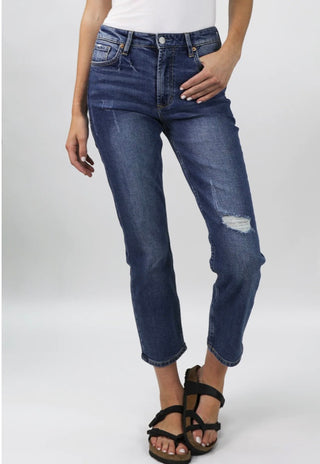 Woman standing in front of white background with hand in front pocket of dark destress denim jeans on the left leg.