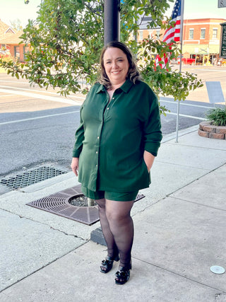 A woman standing on the sidewalk wearing a green button down playsuit with long sleeves, posing with a tree and a flag in the background.