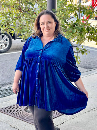A woman standing on the sidewalk wearing a blue velvet 3/4 sleeve dress with a high-low cut, posing with one hand against a tree.