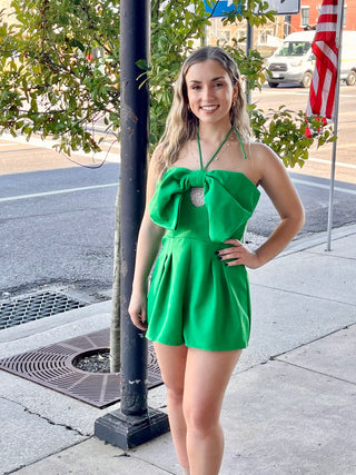 A woman standing on the sidewalk wearing a green sleeveless romper with a bow front design, posing against a backdrop of a city street.