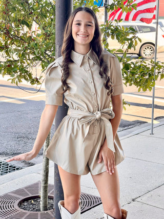 A woman standing on the sidewalk wearing a beige faux leather belted romper with button front, paired with white cowboy boots.