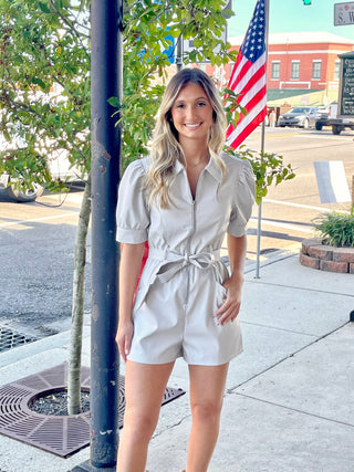 A woman standing by a pole, wearing a light beige short sleeve romper with a belt, pockets, and a zipper front.