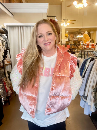 A woman standing in a retail environment wearing a pink shimmer sleeveless button down ruffle shoulder puffer vest.