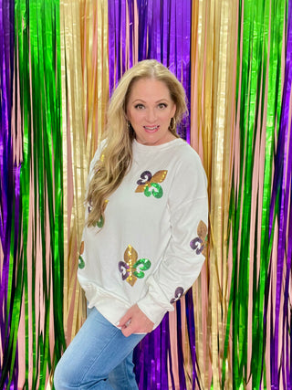 A person standing in front of Mardi Gras backdrop, wearing a white sequin pullover sweatshirt with a fleur-de-lis pattern that are purple, green and gold and a smile on their face.