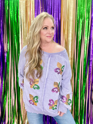 A woman standing in front of a backdrop with green, gold and purple tensile, wearing a purple waffle shirt with purple, green and gold fleur-de-lis patchwork. Shirt is a scoop neck.