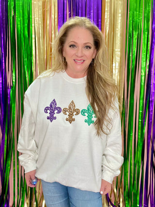 A woman smiling, wearing a white fleur-de-lis and sequin sweatshirt, with green and purple sequins in the background.