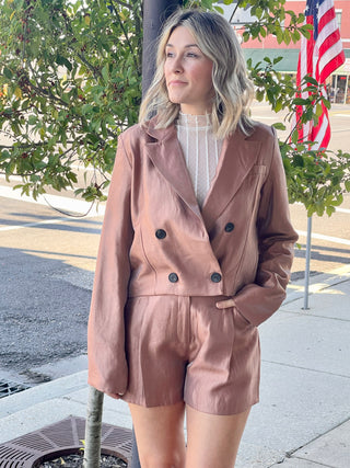 A person standing on the sidewalk wearing high waisted shorts and a jacket, with a background of trees and a flag.