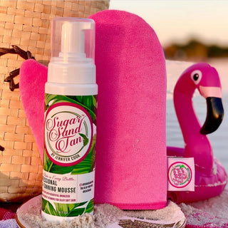 A bottle of Sugar Sand Tan self-tanning mousse is displayed next to a pink tanning mitt and a pink flamingo float, set against a beach-themed backdrop.