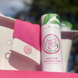 A packaged exfoliating glove with the brand 'Sugar Sand Tan' displayed on the packaging, alongside a pink washcloth, all set against a blurred background.