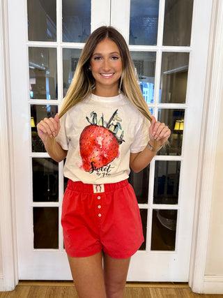 A person wearing a white t-shirt with a red strawberry graphic and red shorts, standing in front of a glass door.
