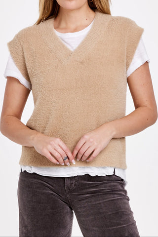 Person wearing a beige sweater vest over a white shirt with dark jeans on a white background
