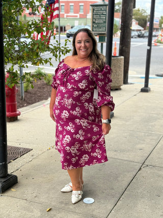 Woman in a purple floral dress standing on a sidewalk with a street and trees in the background.