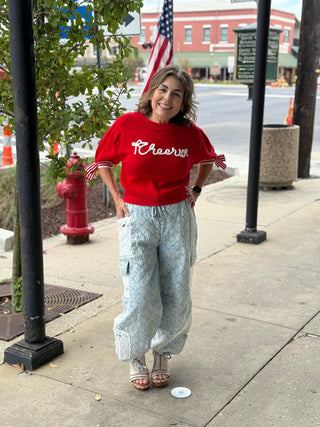 Woman wearing a red 'Cheers' shirt and light blue pants standing on a sidewalk.