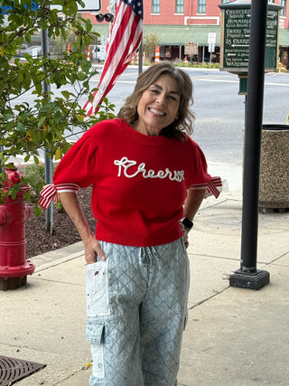 Woman wearing a red 'Cheers' sweater on a sidewalk with an American flag in the background.